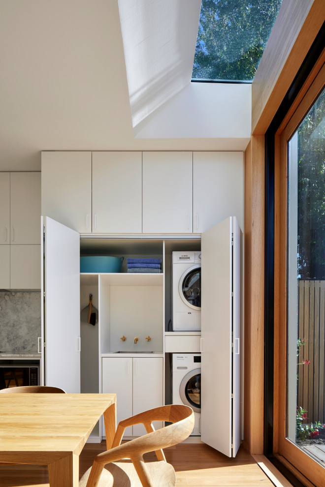 Inspiration for a small contemporary single-wall laundry cupboard in Melbourne with a single-bowl sink, flat-panel cabinets, white cabinets, laminate benchtops, white splashback, white walls, light hardwood floors, a stacked washer and dryer, brown floor and white benchtop.