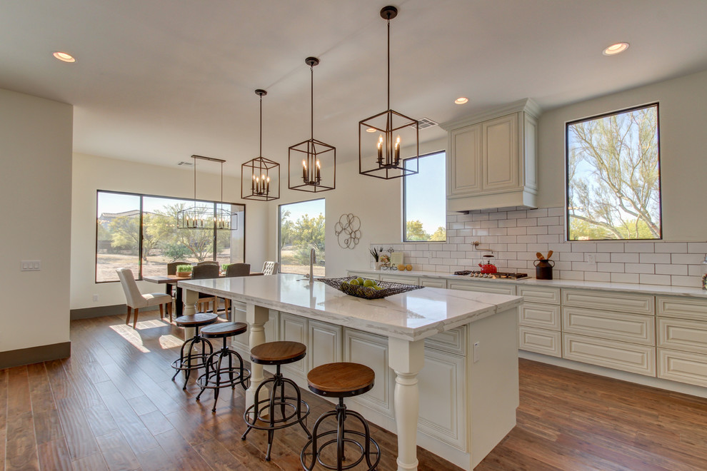 Eat-in kitchen - mid-sized transitional l-shaped dark wood floor and brown floor eat-in kitchen idea in Phoenix with a farmhouse sink, raised-panel cabinets, white cabinets, marble countertops, white backsplash, subway tile backsplash, stainless steel appliances and an island