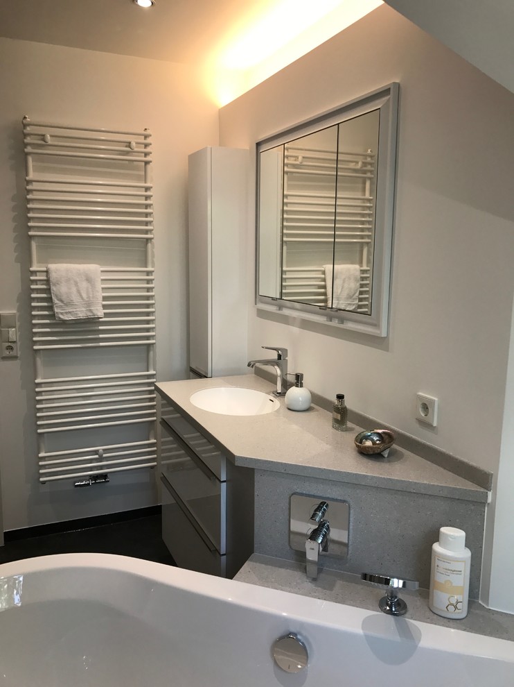 Badezimmer With flächenbündigen Schrankfronten, weißen Schränken, freistehender Badewanne, Wandtoilette, grauen Fliesen, Metallfliesen, weißer Wandfarbe, Vinylboden, Trogwaschbecken, Mineralwerkstoff-Waschtisch and schwarzem Boden in Berlin