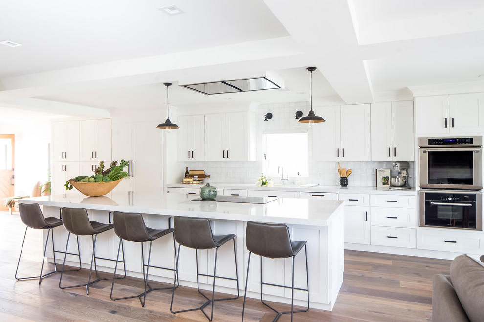 Inspiration for a large transitional galley open plan kitchen in Orange County with a single-bowl sink, shaker cabinets, white cabinets, solid surface benchtops, grey splashback, ceramic splashback, stainless steel appliances, medium hardwood floors, with island and brown floor.