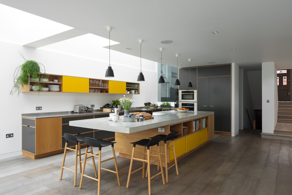 Photo of a large modern l-shaped kitchen in London with concrete benchtops, stainless steel appliances, with island, flat-panel cabinets, yellow cabinets, light hardwood floors, grey floor and grey benchtop.