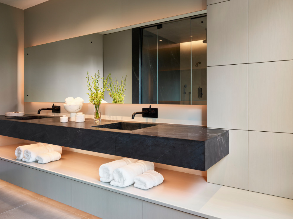 North Shore Less is More - This primary bath features an asymmetrical design combining stained white oak custom cabinetry which is a beautiful compliment to the integrated black soapstone sink, back lit custom expansive mirror, and additional accent lighting under the cabinetry drawers.  The linear design features multiple layers for visual interest and a unique concealed panel which opens to the laundry room for quick deposit of towels and clothing for washing.