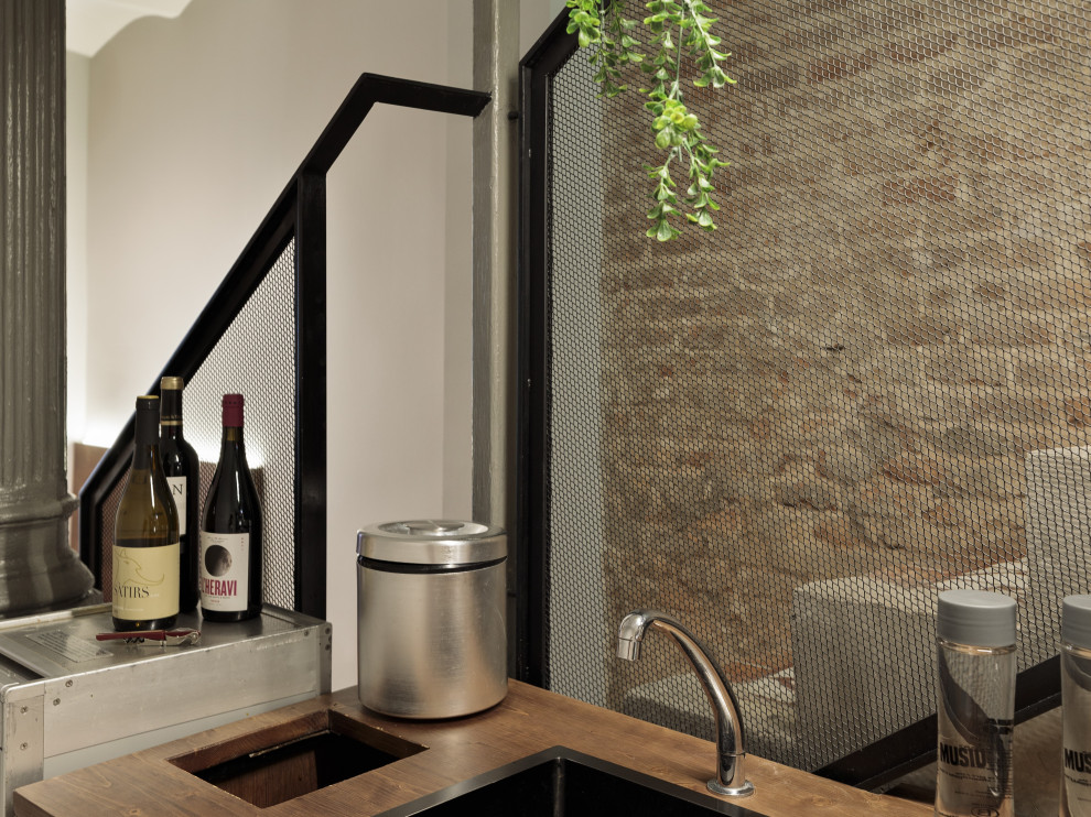 Modelo de bar en casa con barra de bar lineal de tamaño medio con fregadero encastrado, armarios con paneles lisos, puertas de armario de madera en tonos medios, encimera de madera, salpicadero negro, salpicadero de metal, suelo de cemento, suelo gris y encimeras marrones