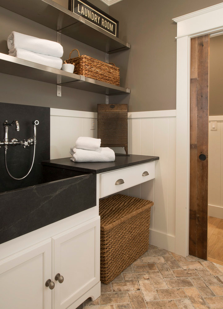 This is an example of a mid-sized traditional galley laundry room in St Louis with flat-panel cabinets, white cabinets, soapstone benchtops, grey walls, brick floors, a side-by-side washer and dryer and a farmhouse sink.
