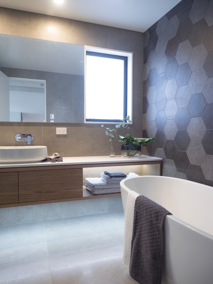 Inspiration for a contemporary bathroom in Auckland with flat-panel cabinets, medium wood cabinets, a freestanding tub, gray tile, grey walls, a vessel sink, grey floor and white benchtops.