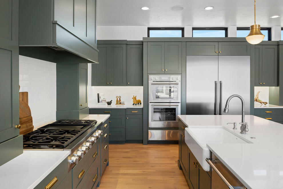 Design ideas for a transitional l-shaped kitchen in Boise with a farmhouse sink, shaker cabinets, green cabinets, white splashback, subway tile splashback, stainless steel appliances, medium hardwood floors, with island, brown floor and white benchtop.