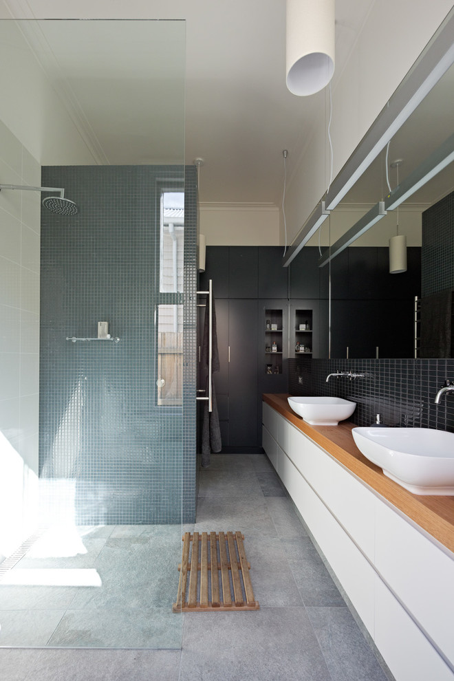 Photo of a contemporary bathroom in Melbourne with a vessel sink, flat-panel cabinets, white cabinets, wood benchtops, a curbless shower, black tile, white walls, concrete floors and brown benchtops.