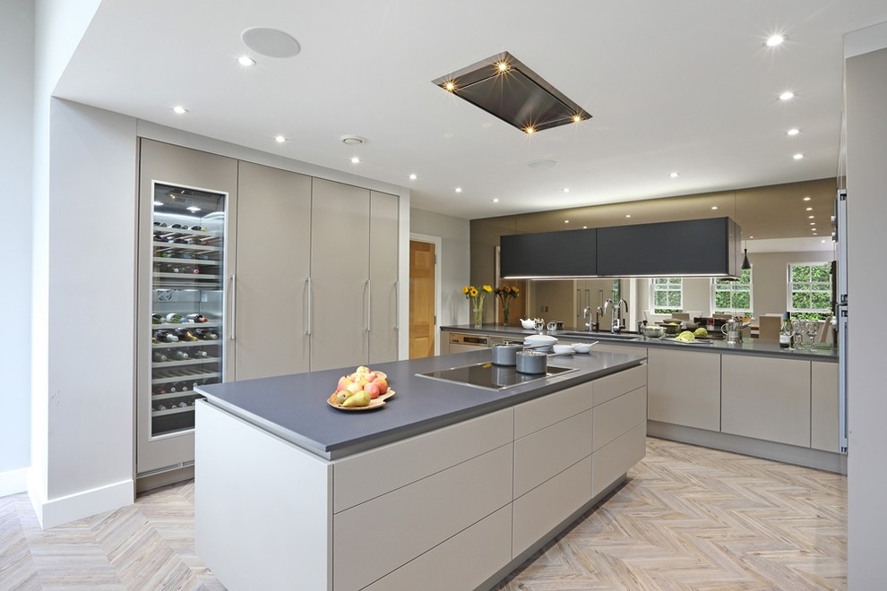 Photo of a large contemporary l-shaped eat-in kitchen in Hertfordshire with an integrated sink, flat-panel cabinets, grey cabinets, solid surface benchtops, metallic splashback, glass sheet splashback, black appliances, light hardwood floors, with island and grey floor.