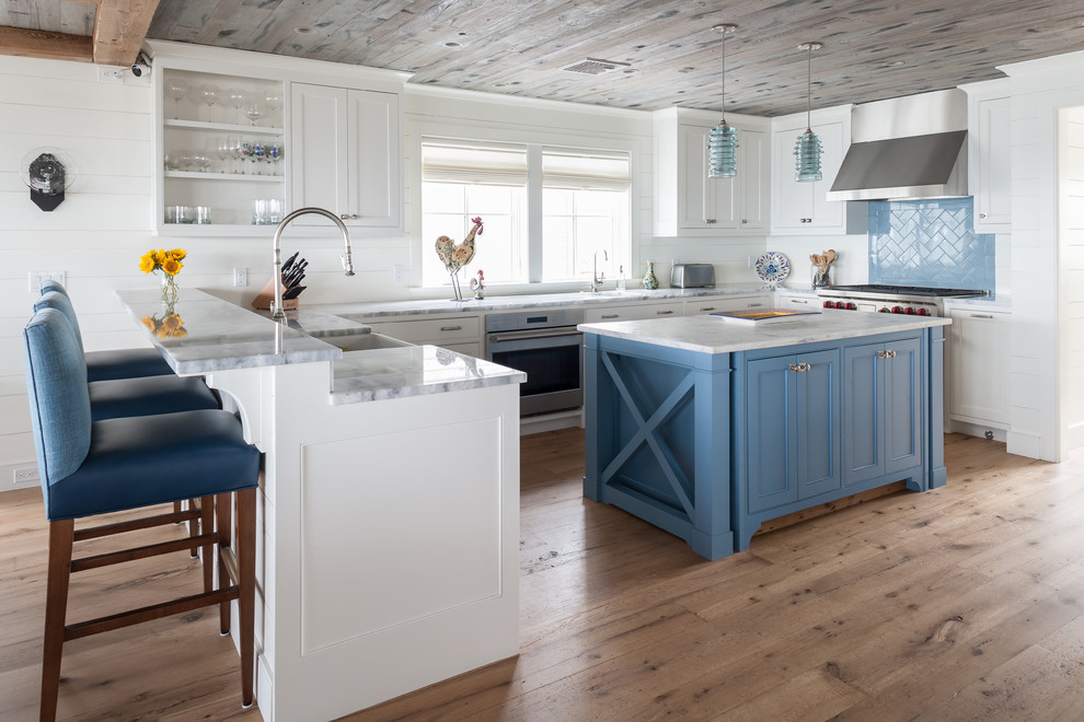 This is an example of a beach style u-shaped kitchen in Bridgeport with a farmhouse sink, recessed-panel cabinets, white cabinets, blue splashback, stainless steel appliances, medium hardwood floors, with island, brown floor and grey benchtop.