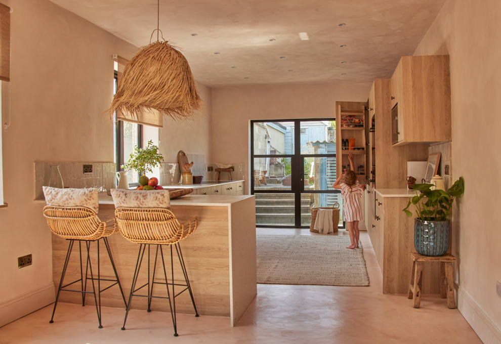 Design ideas for a large mediterranean l-shaped eat-in kitchen in Cornwall with an undermount sink, flat-panel cabinets, light wood cabinets, solid surface benchtops, beige splashback, ceramic splashback, black appliances, concrete floors, a peninsula, pink floor and beige benchtop.