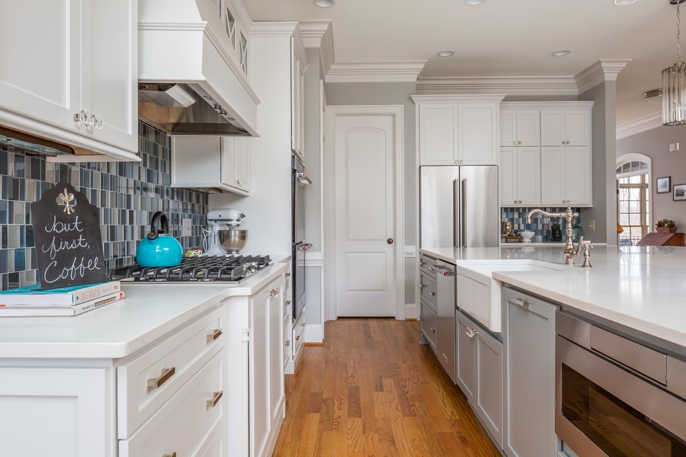 Example of a large transitional l-shaped medium tone wood floor open concept kitchen design in Raleigh with a farmhouse sink, flat-panel cabinets, white cabinets, quartz countertops, blue backsplash, glass tile backsplash, stainless steel appliances and an island