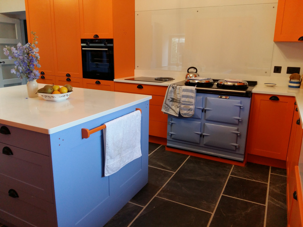 This is an example of a large contemporary u-shaped open plan kitchen in Cornwall with a farmhouse sink, shaker cabinets, orange cabinets, quartz benchtops, white splashback, black appliances, slate floors, grey floor, white benchtop and exposed beam.