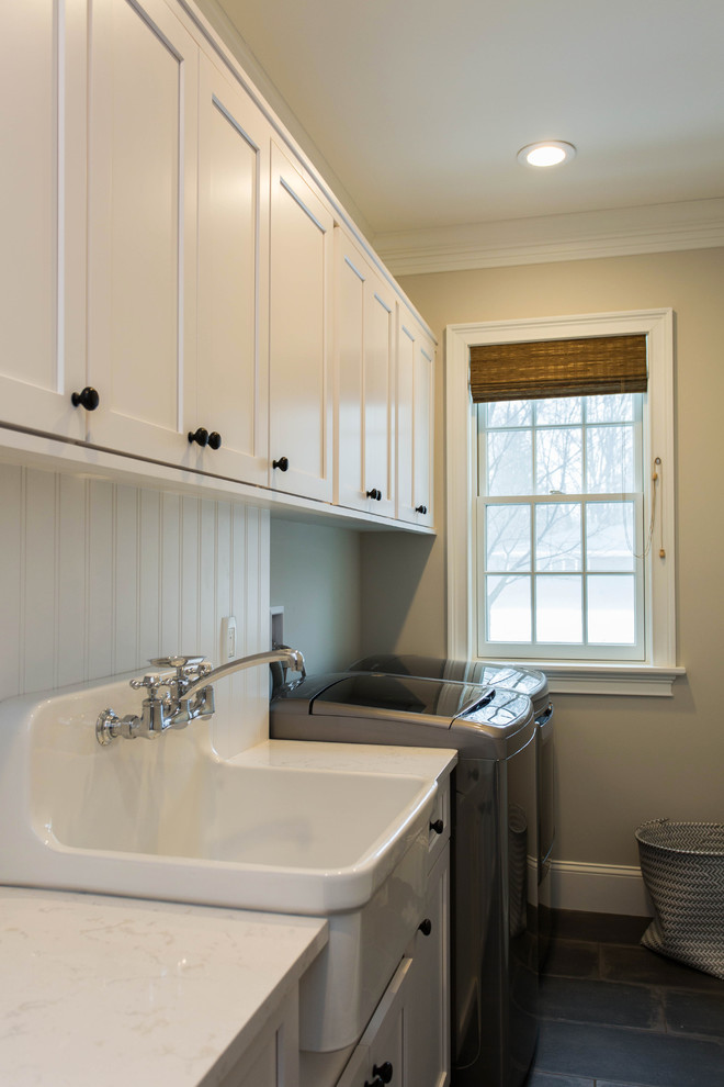 This is an example of a mid-sized transitional single-wall dedicated laundry room in Boston with recessed-panel cabinets, white cabinets, quartz benchtops, a side-by-side washer and dryer, grey floor, a farmhouse sink, beige walls and concrete floors.
