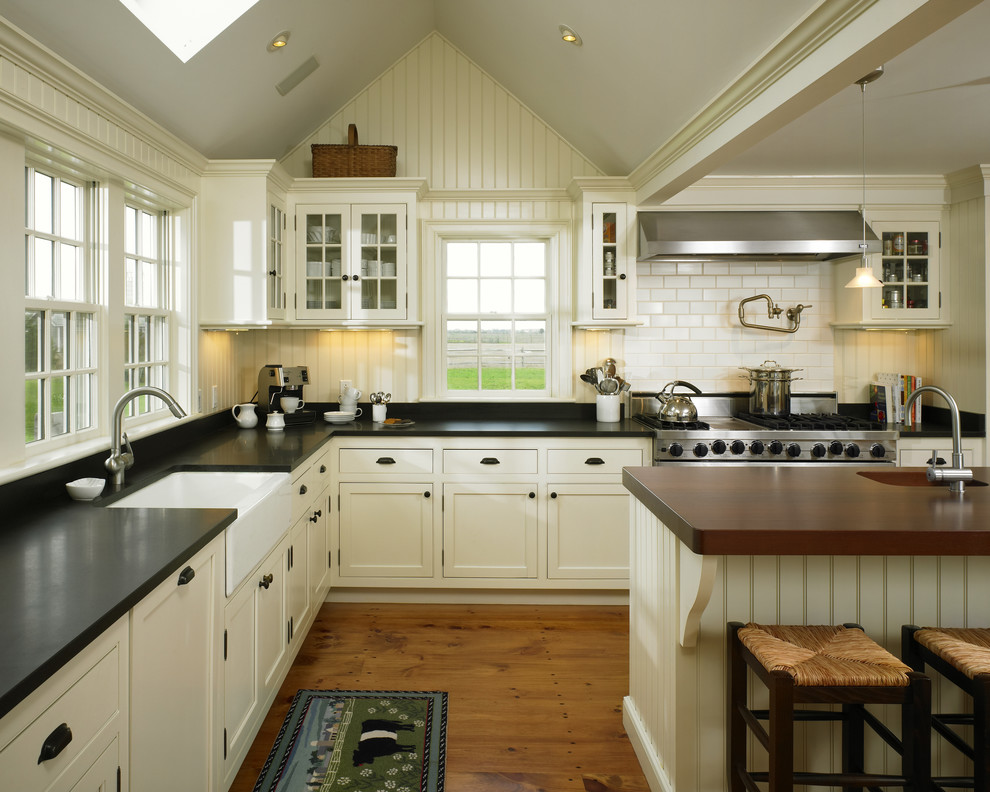 Design ideas for a large country l-shaped open plan kitchen in Boston with a farmhouse sink, glass-front cabinets, white cabinets, white splashback, subway tile splashback, stainless steel appliances, medium hardwood floors, with island and black benchtop.