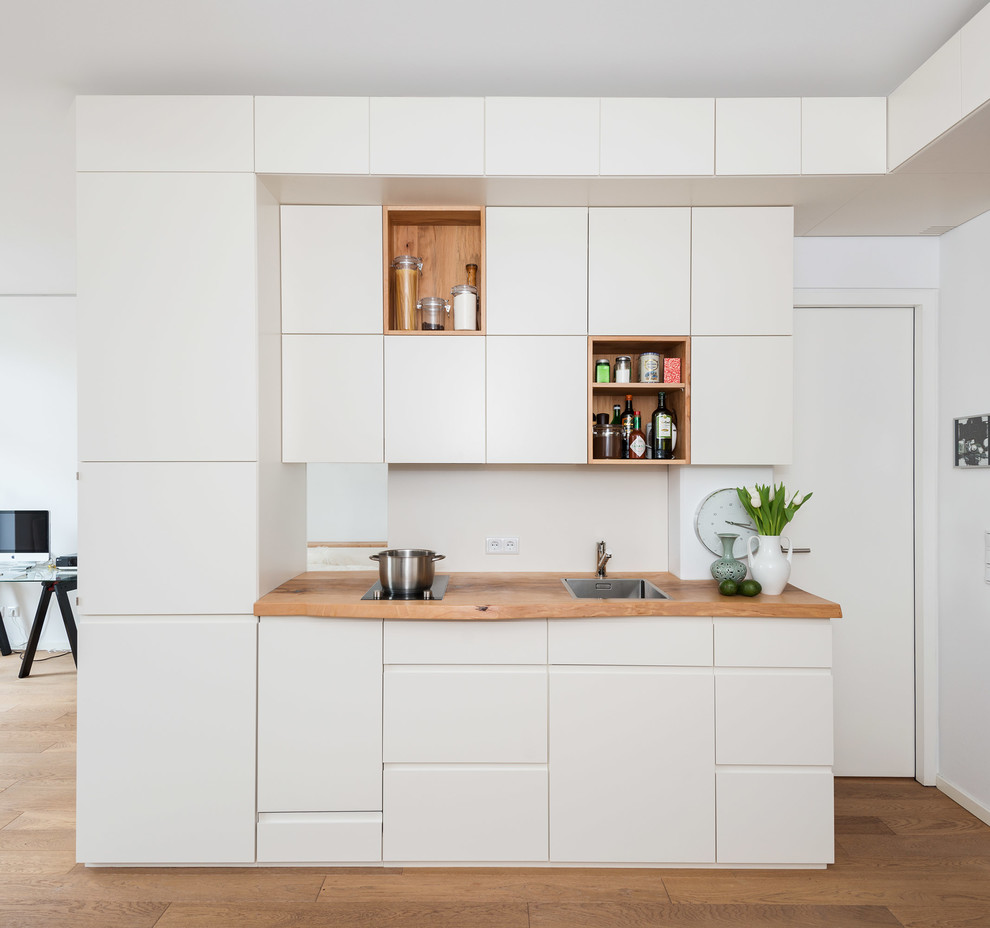 Photo of a small contemporary single-wall open plan kitchen in Berlin with flat-panel cabinets, white cabinets, wood benchtops, medium hardwood floors, a drop-in sink and white splashback.