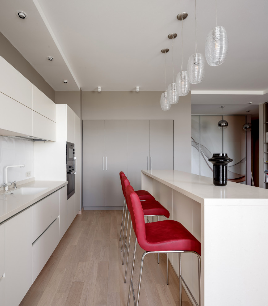 Mid-sized contemporary l-shaped open plan kitchen in Other with flat-panel cabinets, white cabinets, quartz benchtops, white splashback, porcelain splashback, black appliances, with island, beige benchtop, an undermount sink, light hardwood floors and beige floor.