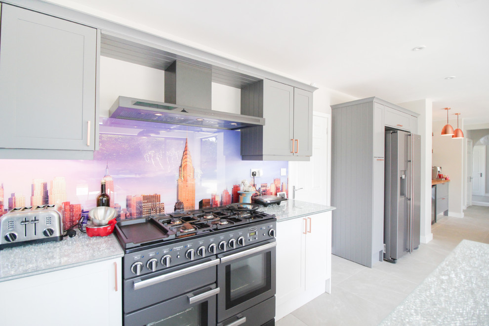 This is an example of an expansive contemporary u-shaped open plan kitchen in London with an integrated sink, shaker cabinets, white cabinets, glass benchtops, multi-coloured splashback, glass sheet splashback, black appliances, ceramic floors, with island, grey floor and green benchtop.