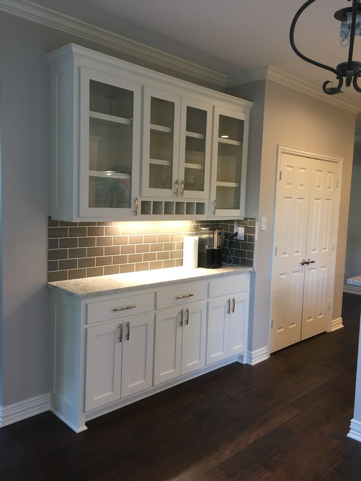 Small traditional single-wall wet bar in Dallas with white cabinets, granite benchtops, grey splashback, glass tile splashback, dark hardwood floors, brown floor and glass-front cabinets.