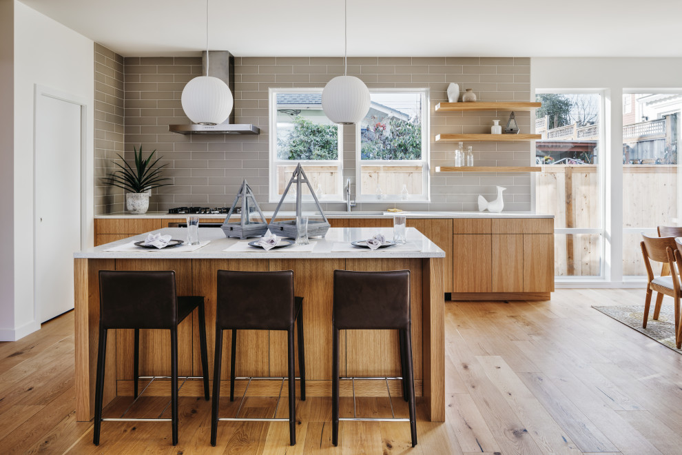 Photo of a mid-sized modern single-wall open plan kitchen in Los Angeles with an undermount sink, flat-panel cabinets, light wood cabinets, quartz benchtops, grey splashback, subway tile splashback, stainless steel appliances, light hardwood floors, with island, beige floor and white benchtop.