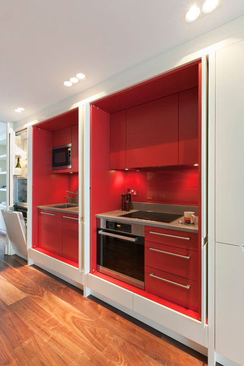 Concealed kitchenette with glossy red slab kitchen cabinets behind pocket doors, integrated sink, cooktop, oven, and microwave