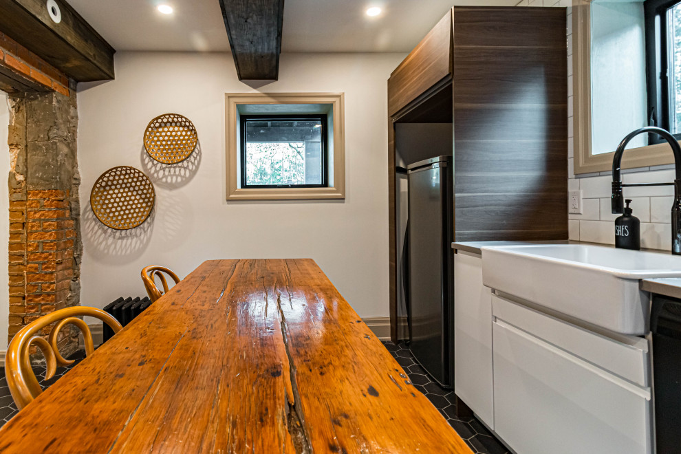 Example of a mid-sized mountain style u-shaped ceramic tile, black floor and exposed beam eat-in kitchen design in Toronto with a farmhouse sink, flat-panel cabinets, white cabinets, quartz countertops, white backsplash, subway tile backsplash, stainless steel appliances, an island and gray countertops