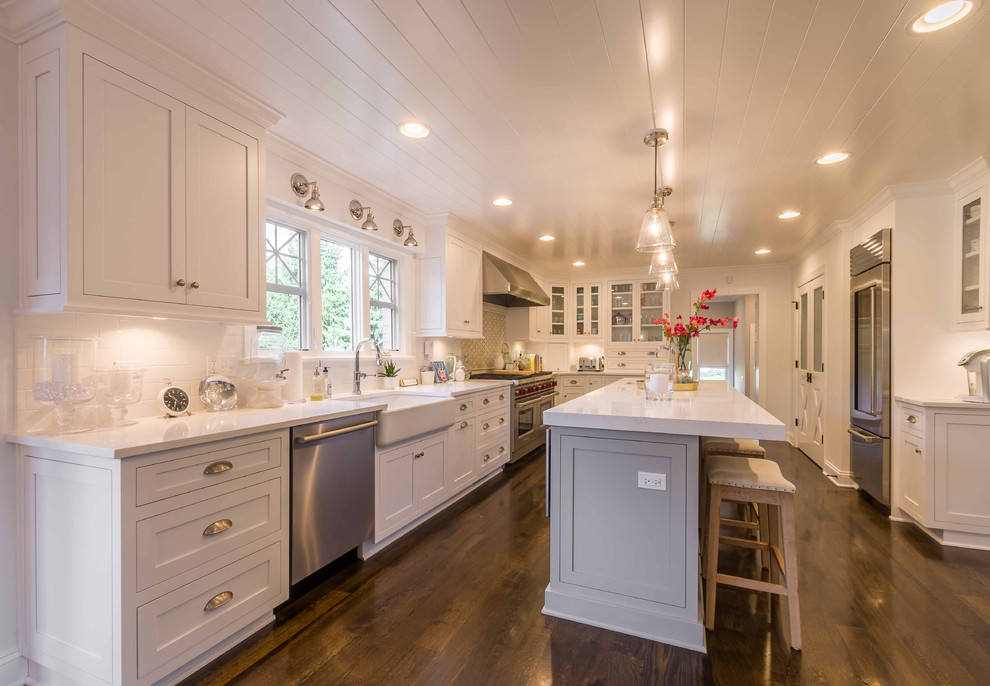 Large transitional l-shaped eat-in kitchen in Chicago with a farmhouse sink, shaker cabinets, white cabinets, marble benchtops, white splashback, subway tile splashback, stainless steel appliances, dark hardwood floors, with island, brown floor, white benchtop and timber.