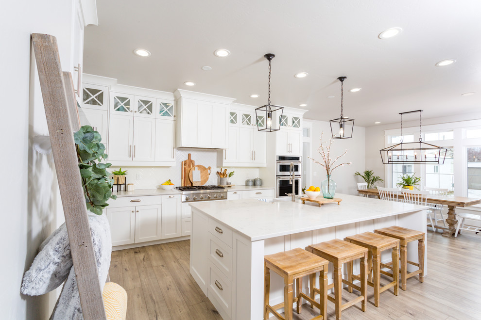 Large beach style eat-in kitchen in San Francisco with a farmhouse sink, shaker cabinets, white cabinets, quartz benchtops, white splashback, stainless steel appliances, light hardwood floors, with island, beige floor and white benchtop.