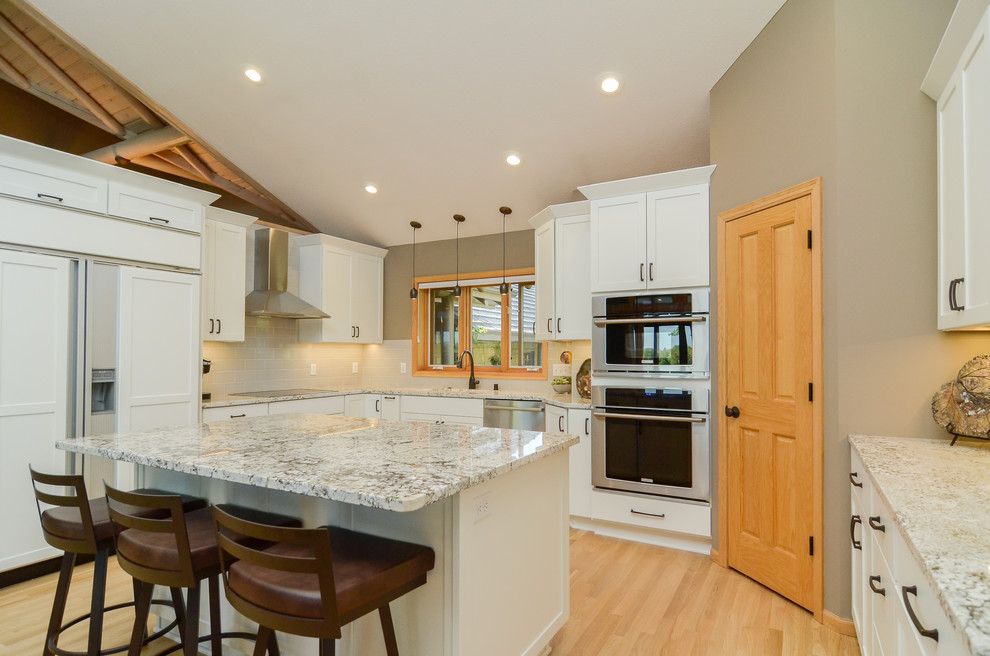Large transitional u-shaped kitchen in Minneapolis with an undermount sink, white cabinets, granite benchtops, grey splashback, subway tile splashback, stainless steel appliances, with island, brown floor, shaker cabinets, light hardwood floors and multi-coloured benchtop.