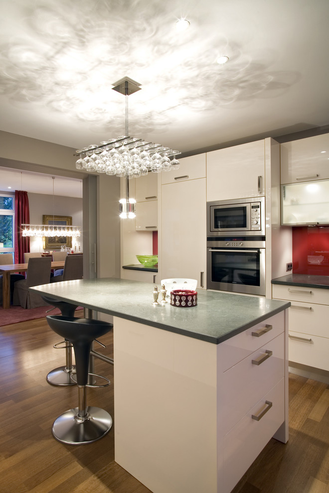 Mid-sized contemporary single-wall open plan kitchen in Cologne with flat-panel cabinets, white cabinets, red splashback, glass sheet splashback, stainless steel appliances, medium hardwood floors and with island.