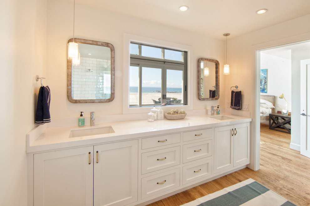 Photo of a mid-sized beach style master bathroom in Orange County with an undermount sink, beaded inset cabinets, white cabinets, an alcove shower, white tile, subway tile, white walls, light hardwood floors, solid surface benchtops and white benchtops.