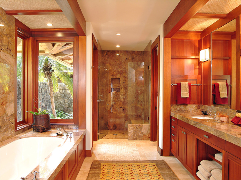 Photo of a mid-sized tropical master bathroom in Hawaii with an undermount sink, shaker cabinets, medium wood cabinets, an undermount tub, an alcove shower, brown tile, stone slab, white walls, travertine floors, beige floor, a hinged shower door and grey benchtops.
