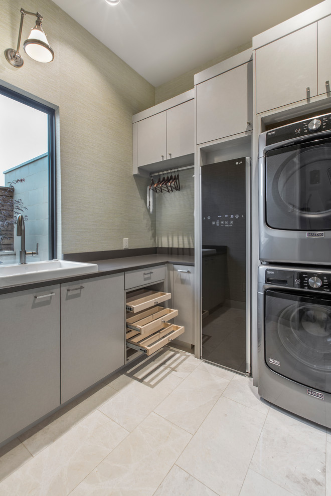 Inspiration for a small contemporary l-shaped dedicated laundry room in Dallas with a drop-in sink, flat-panel cabinets, a stacked washer and dryer, beige floor, grey cabinets, grey walls and black benchtop.