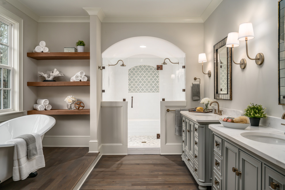 This is an example of a transitional master bathroom in Charlotte with recessed-panel cabinets, grey cabinets, a freestanding tub, a double shower, grey walls, dark hardwood floors, an undermount sink, brown floor, a hinged shower door and beige benchtops.