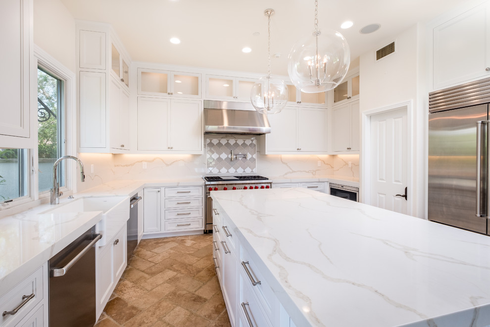 Example of a mid-sized transitional l-shaped porcelain tile and beige floor enclosed kitchen design in Orange County with a farmhouse sink, recessed-panel cabinets, white cabinets, quartz countertops, white backsplash, marble backsplash, stainless steel appliances and an island
