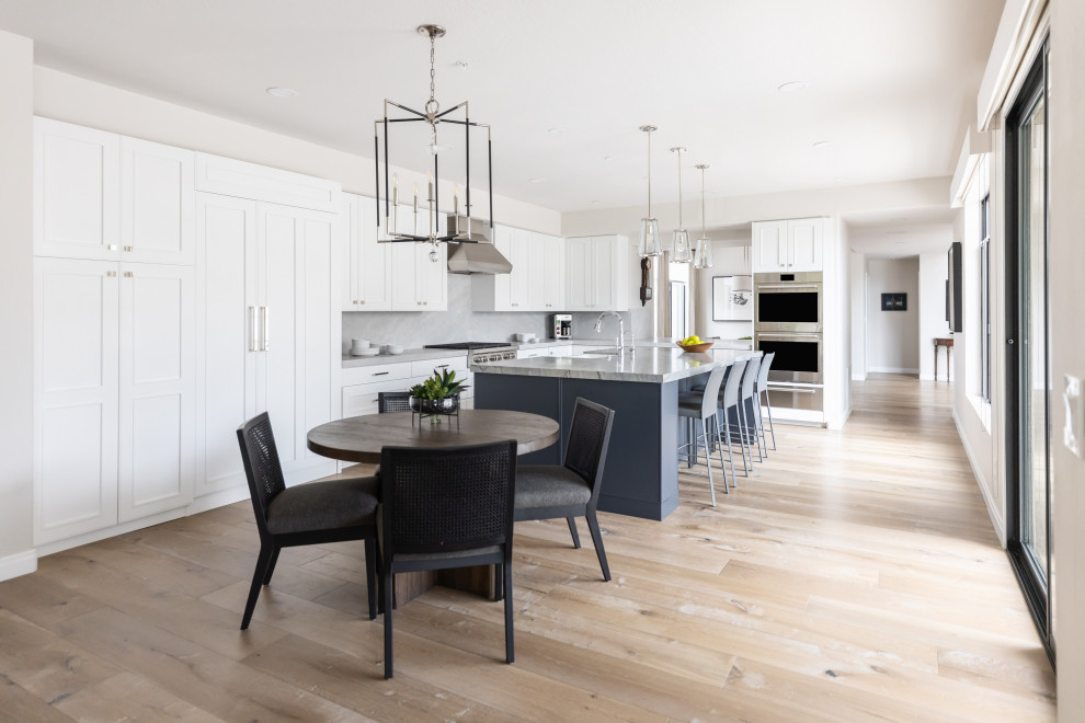 Eat-in kitchen - large transitional medium tone wood floor and brown floor eat-in kitchen idea in Phoenix with a farmhouse sink, recessed-panel cabinets, white cabinets, quartzite countertops, beige backsplash, stone slab backsplash, paneled appliances, an island and gray countertops