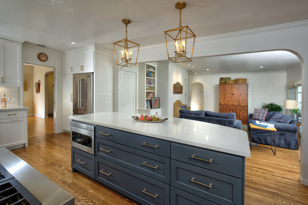 Inspiration for a mid-sized transitional l-shaped open plan kitchen in Sacramento with shaker cabinets, with island, a farmhouse sink, white cabinets, quartz benchtops, white splashback, stone slab splashback, stainless steel appliances, light hardwood floors, brown floor and white benchtop.