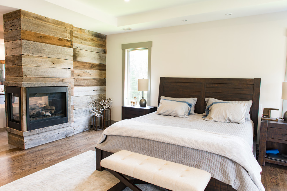This is an example of a large country master bedroom in Charleston with beige walls, dark hardwood floors, a two-sided fireplace, a wood fireplace surround and brown floor.