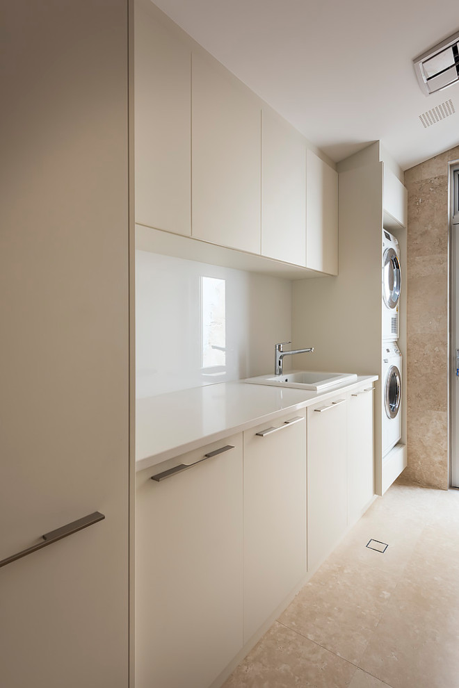Photo of a medium sized modern galley separated utility room in Perth with an utility sink, flat-panel cabinets, beige cabinets, engineered stone countertops, white splashback, engineered quartz splashback, beige walls, travertine flooring, a stacked washer and dryer, beige floors, white worktops and a vaulted ceiling.