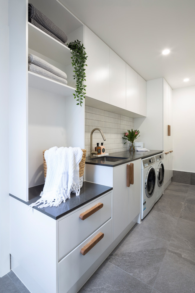 Exempel på en modern svarta linjär svart tvättstuga enbart för tvätt, med en nedsänkt diskho, vita skåp, bänkskiva i kvarts, vitt stänkskydd, stänkskydd i mosaik, vita väggar, klinkergolv i porslin, en tvättmaskin och torktumlare bredvid varandra och grått golv