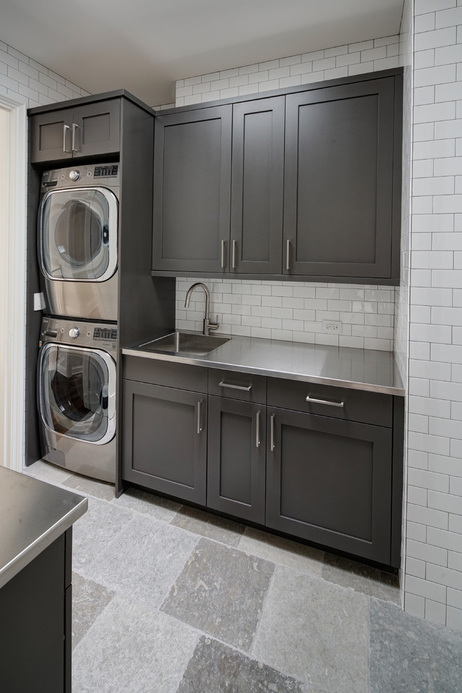 Transitional galley laundry room in Chicago with an integrated sink, recessed-panel cabinets, grey cabinets, stainless steel benchtops, white walls, porcelain floors, a stacked washer and dryer and grey floor.