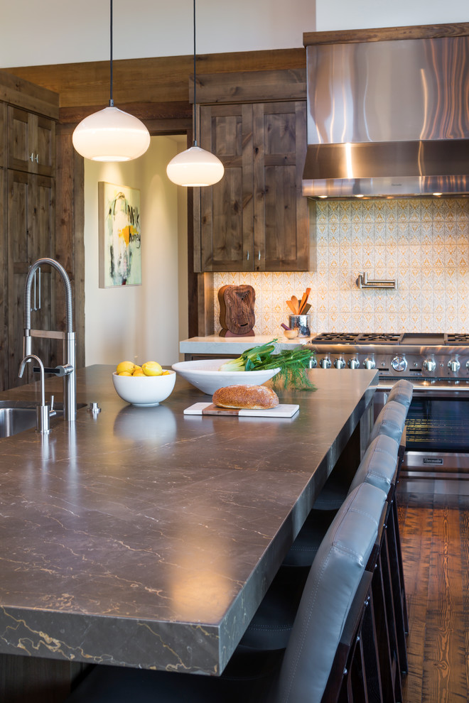 This is an example of a large country l-shaped eat-in kitchen in Other with an undermount sink, shaker cabinets, brown cabinets, limestone benchtops, yellow splashback, terra-cotta splashback, stainless steel appliances, dark hardwood floors, with island and brown floor.