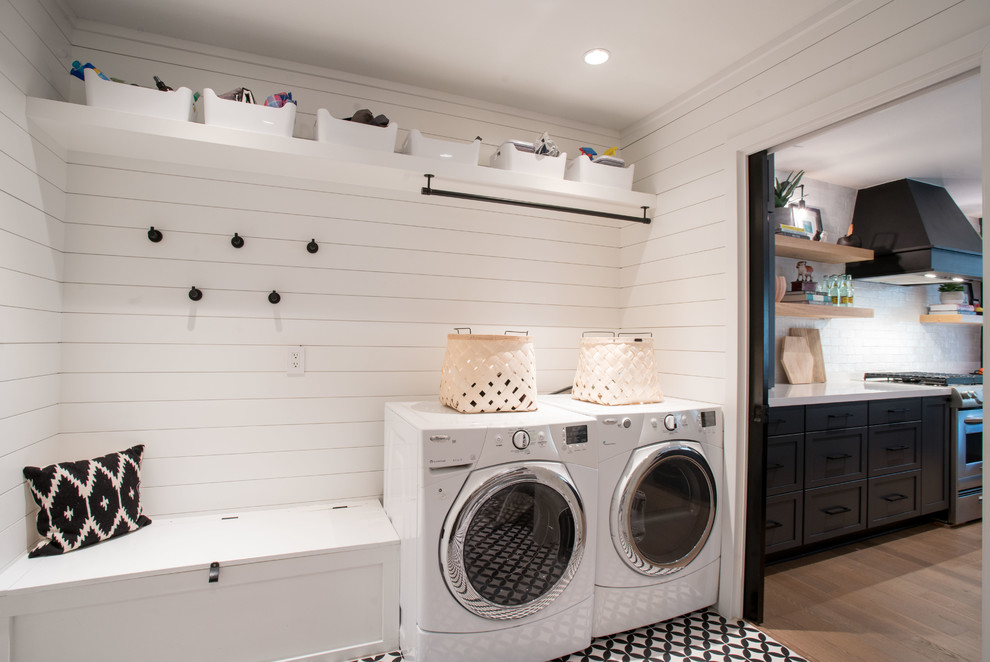 Photo of a mid-sized country dedicated laundry room in Austin with shaker cabinets, white cabinets, white walls, concrete floors, a side-by-side washer and dryer, black floor, white splashback, shiplap splashback and wood walls.
