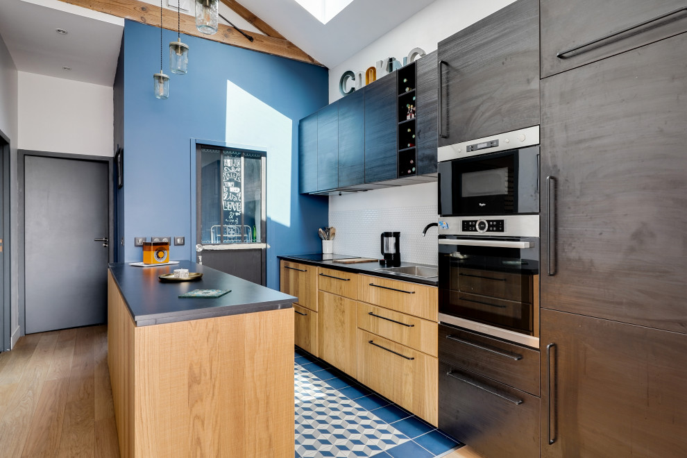 Industrial galley kitchen in Bordeaux with a drop-in sink, flat-panel cabinets, light wood cabinets, stainless steel appliances, a peninsula, blue floor and black benchtop.