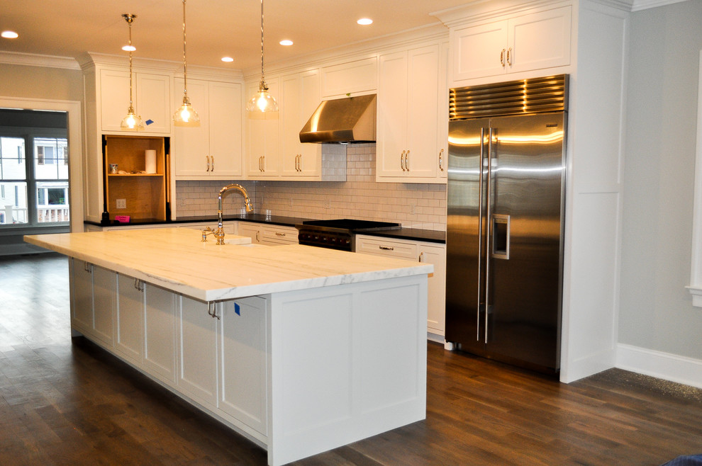 This is an example of a large transitional l-shaped eat-in kitchen in New York with a farmhouse sink, shaker cabinets, white cabinets, soapstone benchtops, white splashback, ceramic splashback, stainless steel appliances, dark hardwood floors, with island, multi-coloured floor and black benchtop.