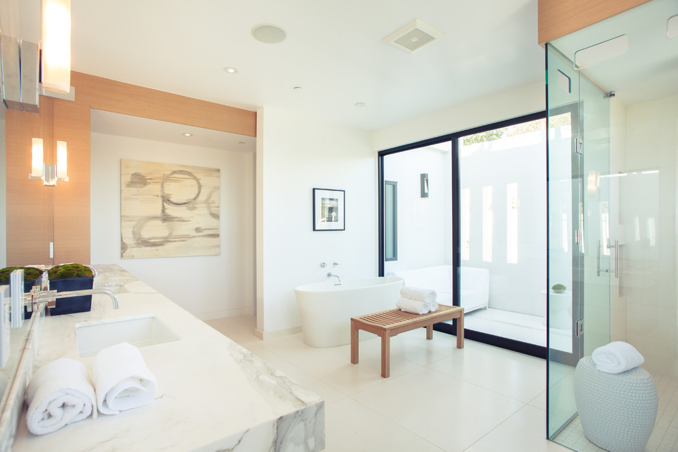 This is an example of an expansive contemporary master bathroom in Los Angeles with an undermount sink, flat-panel cabinets, medium wood cabinets, marble benchtops, a freestanding tub, a corner shower, a one-piece toilet, beige tile, porcelain tile, white walls and limestone floors.