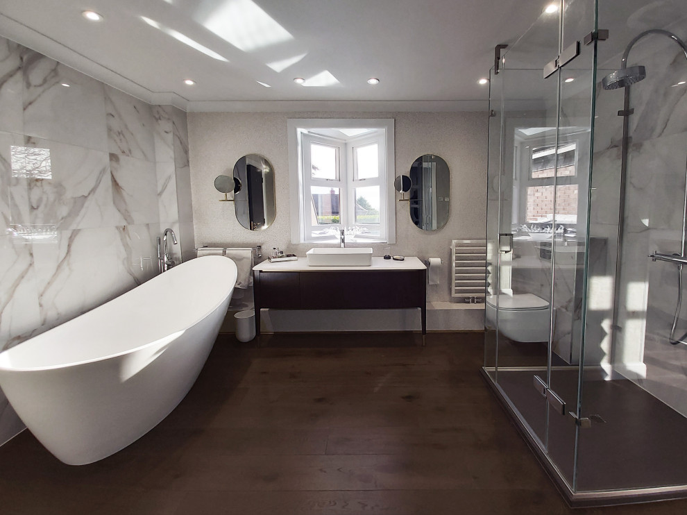 Photo of a mid-sized contemporary master bathroom with flat-panel cabinets, dark wood cabinets, a freestanding tub, a wall-mount toilet, beige tile, marble, beige walls, dark hardwood floors, quartzite benchtops, a hinged shower door and white benchtops.