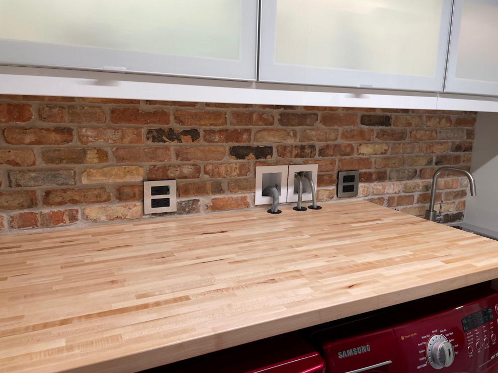 Mid-sized mid-century modern galley concrete floor and white floor dedicated laundry room photo in Chicago with a drop-in sink, glass-front cabinets, white cabinets, wood countertops, brick backsplash and a side-by-side washer/dryer