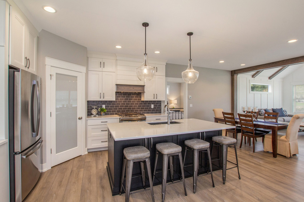Photo of a mid-sized country l-shaped open plan kitchen in Grand Rapids with a single-bowl sink, shaker cabinets, black cabinets, quartz benchtops, black splashback, ceramic splashback, stainless steel appliances, vinyl floors, with island, brown floor and white benchtop.