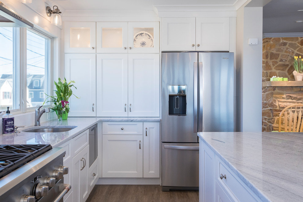This is an example of a mid-sized beach style u-shaped eat-in kitchen in New York with an undermount sink, shaker cabinets, white cabinets, quartzite benchtops, grey splashback, marble splashback, stainless steel appliances, painted wood floors, with island, brown floor and white benchtop.