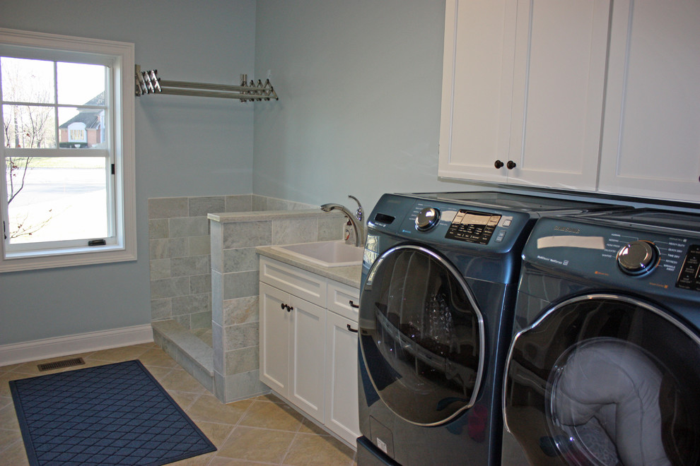 Large arts and crafts single-wall utility room in Chicago with a drop-in sink, recessed-panel cabinets, white cabinets, granite benchtops, porcelain floors, a concealed washer and dryer, beige floor and grey walls.